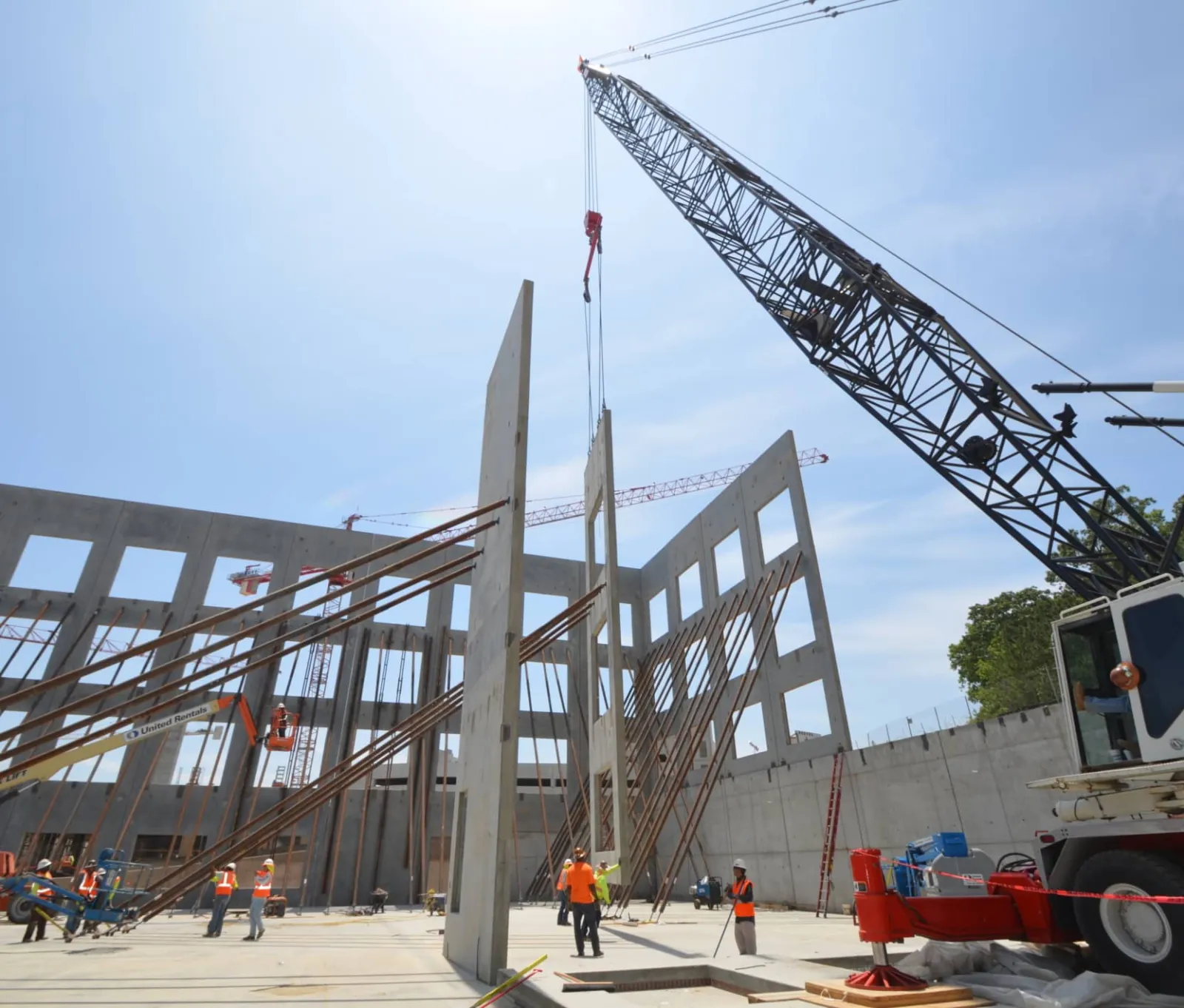 Construction site with workers and crane lifting large concrete wall panels into place under clear sky.