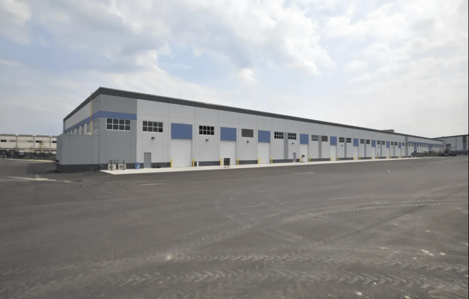 Large industrial warehouse with multiple loading dock doors under a cloudy sky and empty asphalt yard.