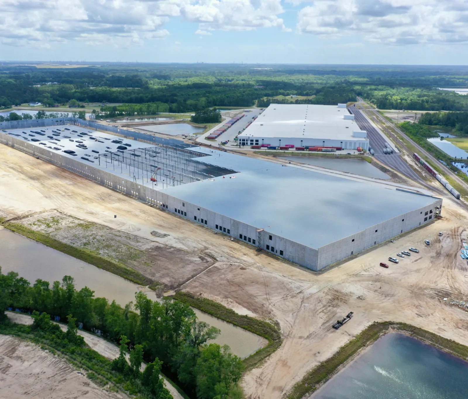 Aerial view of large industrial warehouse buildings surrounded by construction and water bodies under a partly cloudy sky.