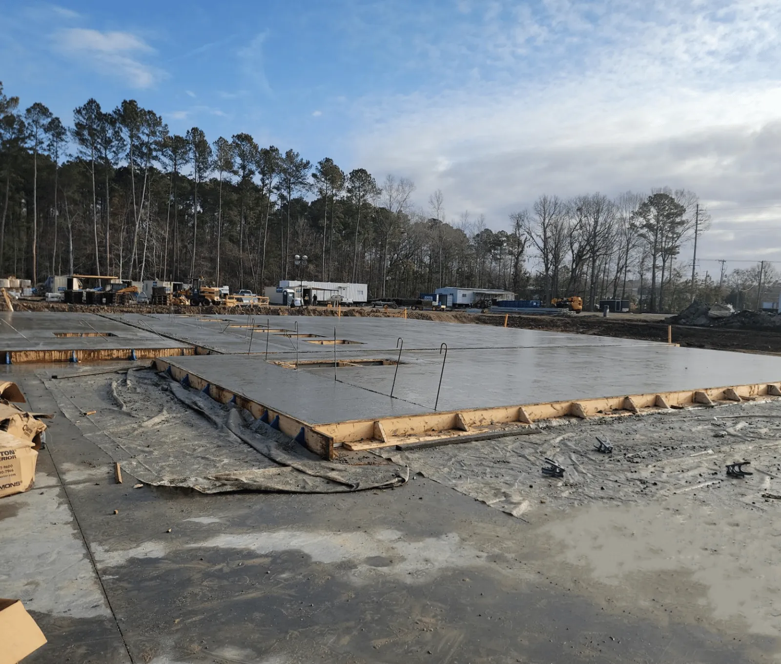 Freshly poured concrete slab foundation at a construction site surrounded by trees and equipment under cloudy sky