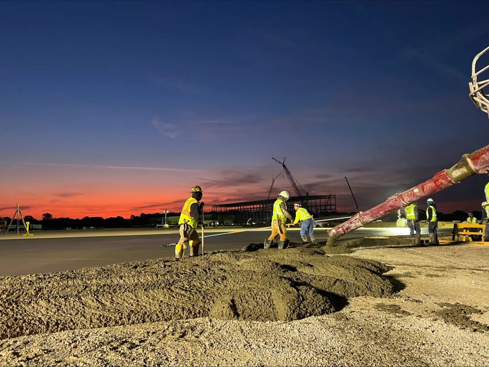 Construction workers pour and level concrete at a site during sunset with cranes visible in the background.