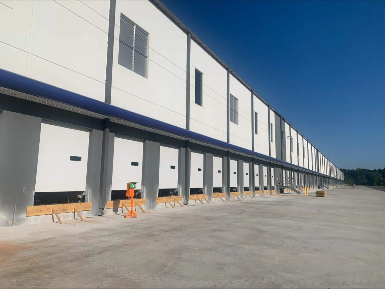 Long row of loading dock doors on a large white warehouse building under clear blue sky.