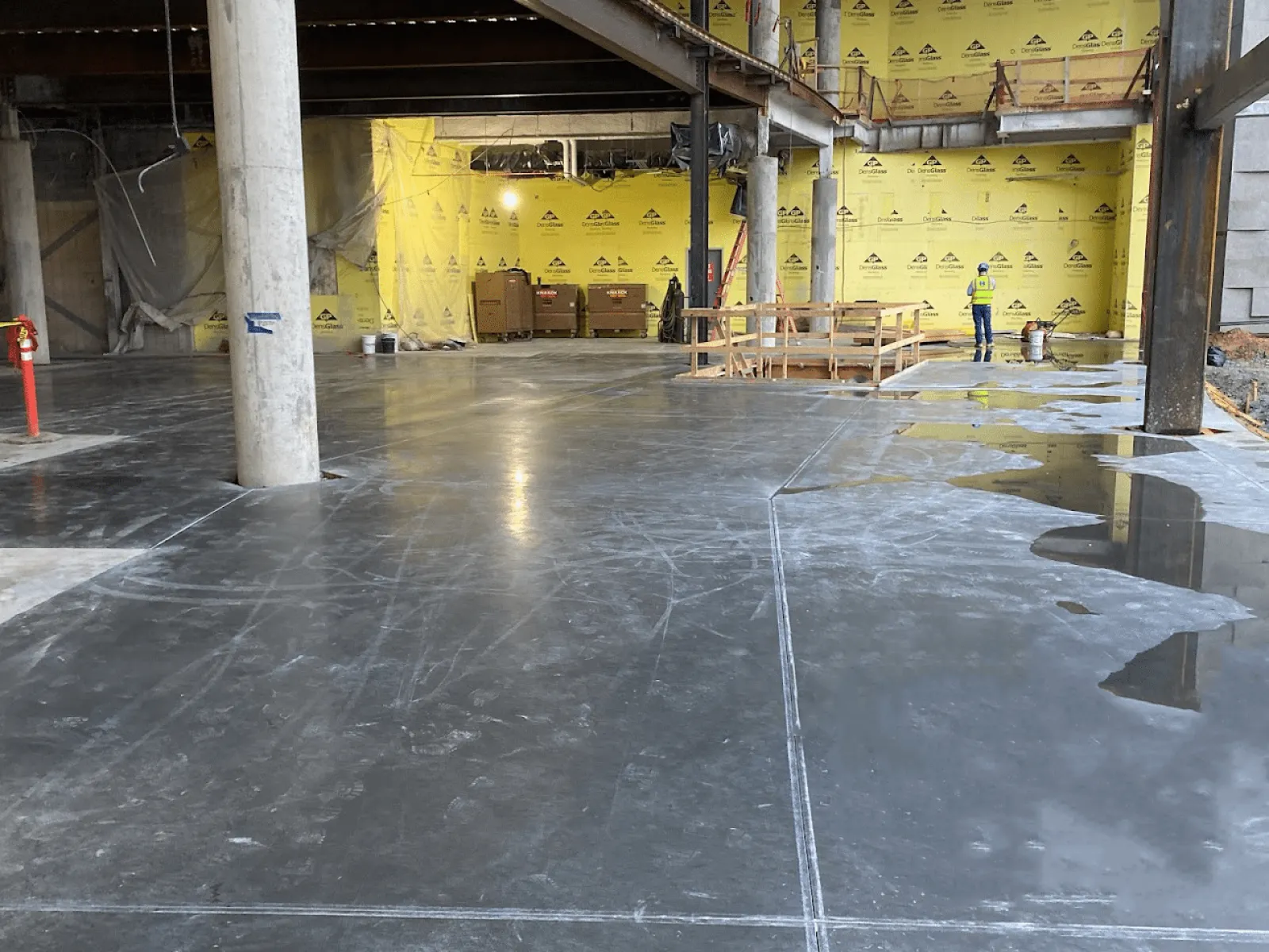 Interior construction site with polished concrete floor, yellow insulation walls, and worker in safety gear.
