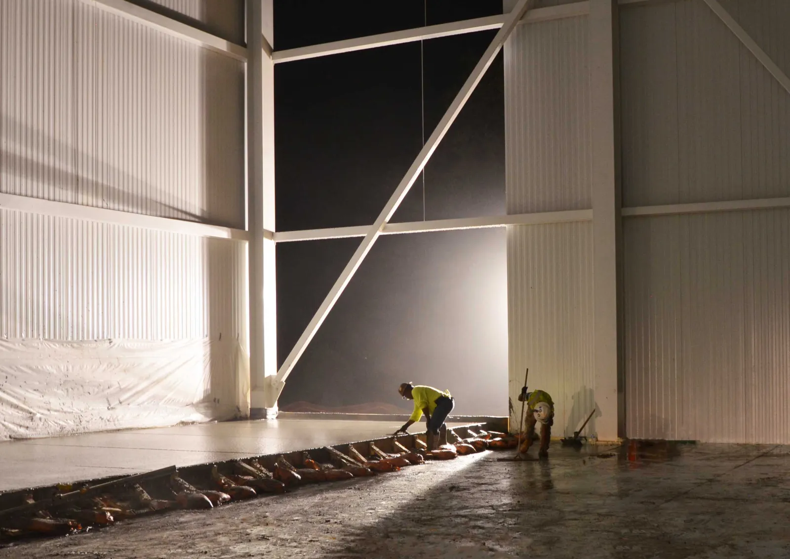 Construction workers smoothing fresh concrete floor inside a large industrial building at night