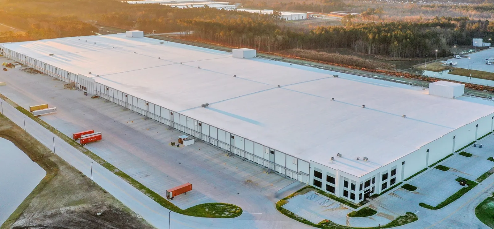 Aerial view of a large white warehouse building with loading docks and trucks at sunset.
