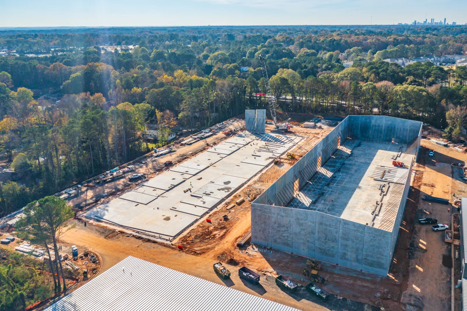 Aerial view of large concrete foundations and walls at a construction site surrounded by trees and city skyline.