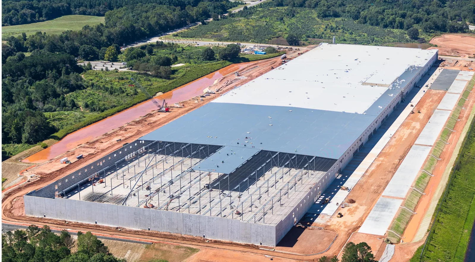 Aerial view of a large warehouse under construction with steel framework and surrounding green landscape.