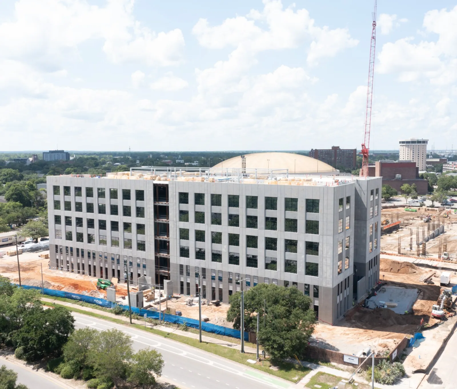 Modern six-story office building under construction with crane and surrounding greenery on a sunny day.