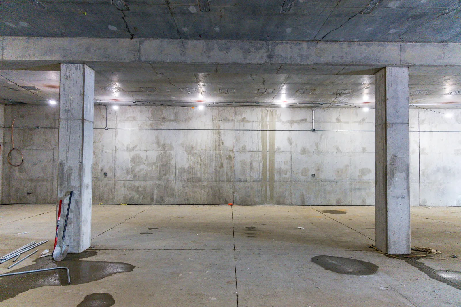 Empty concrete basement with pillars, water puddles, and construction tools scattered on the floor.