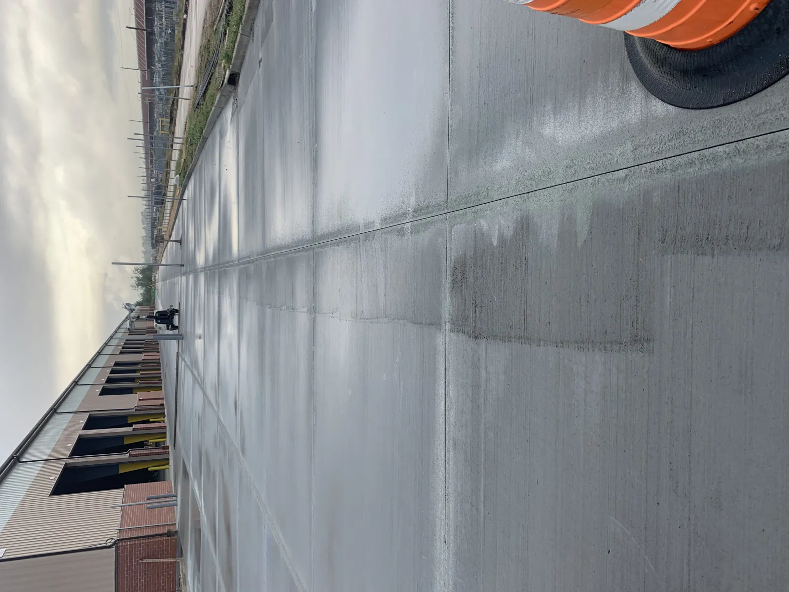 Freshly poured concrete pavement next to industrial building under cloudy sky with orange traffic barrel.