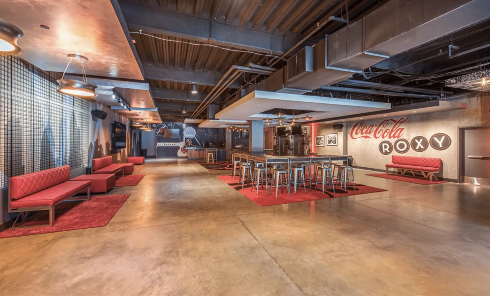 Spacious modern lounge area with red seating, industrial ceiling, bar stools, and Coca-Cola Roxy signage on wall.