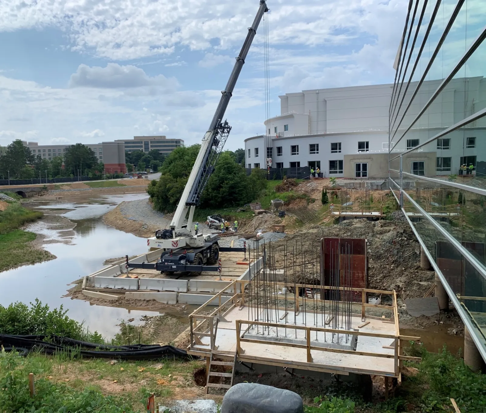 Crane and construction site by a river with steel reinforcements and workers near a large modern building.