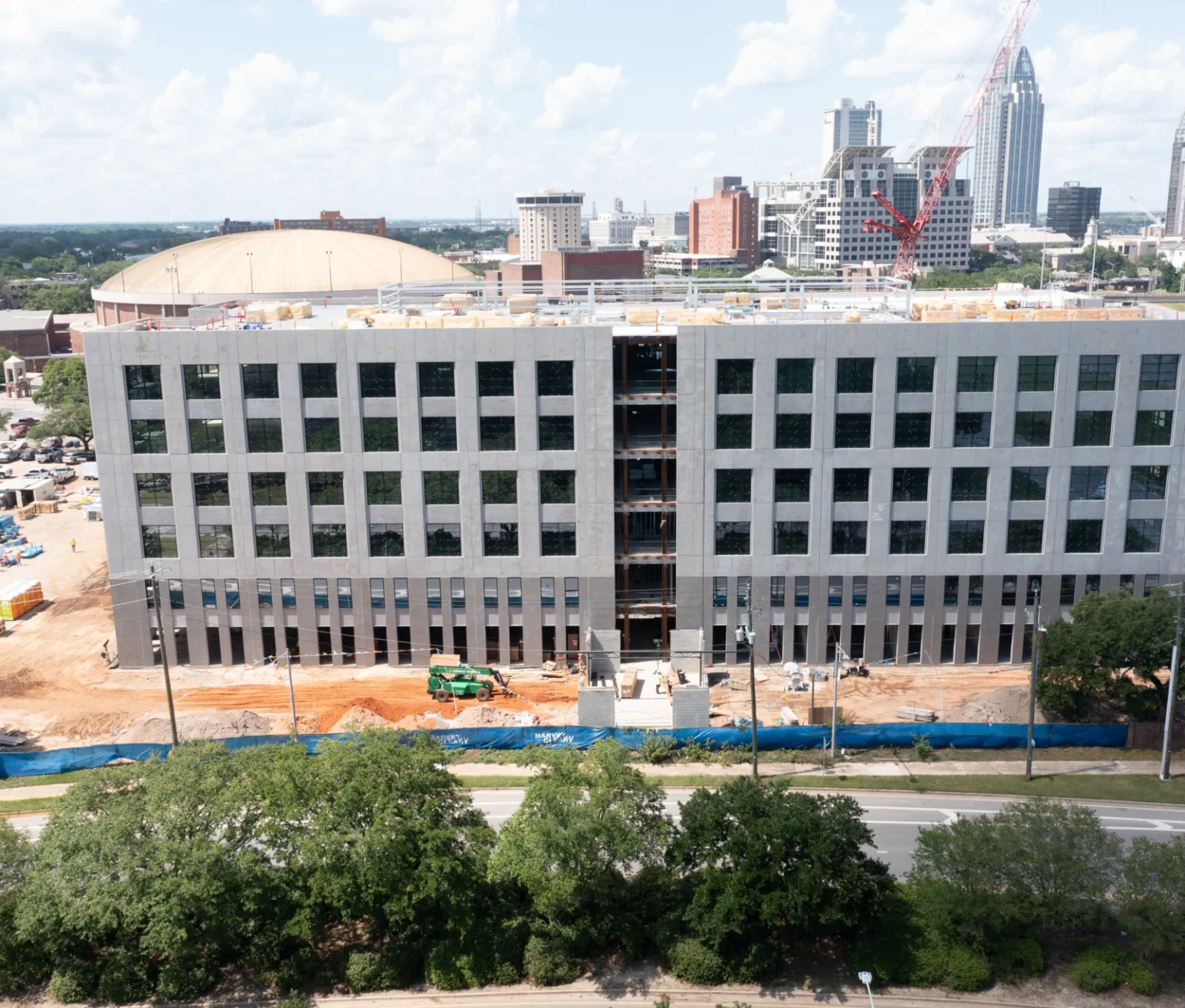 Construction site of a large multi-story building with city skyline and trees in the background under blue sky