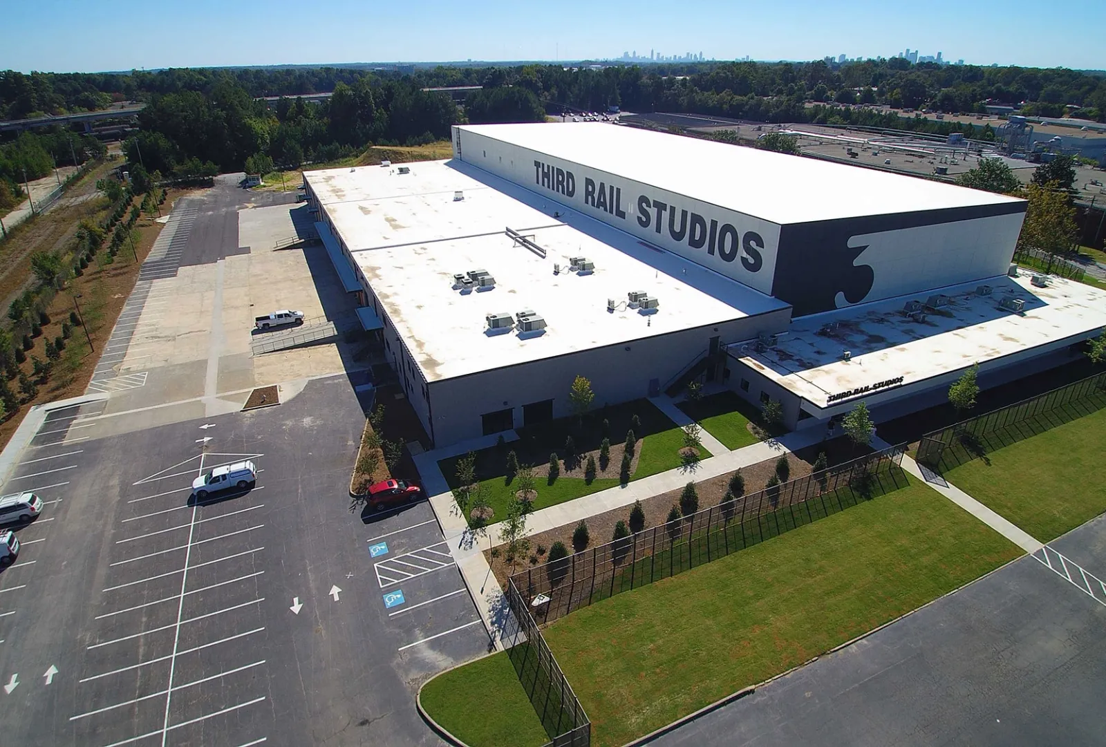 Aerial view of Third Rail Studios building with parking lot, greenery, and city skyline in the distance on a clear day