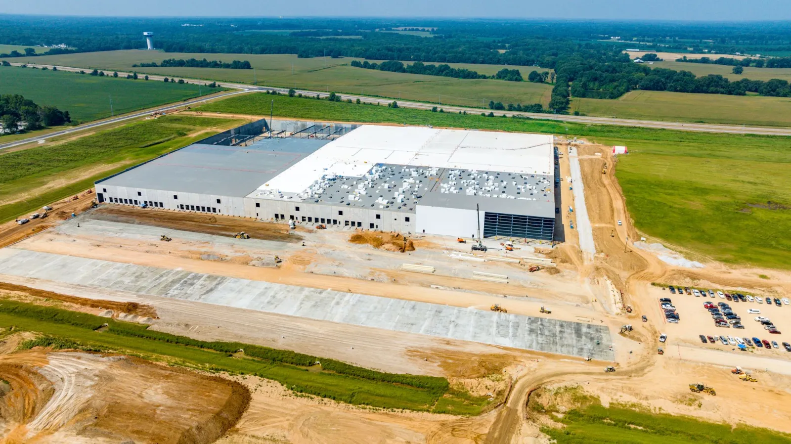Aerial view of a large industrial warehouse under construction surrounded by fields and a dirt parking area.