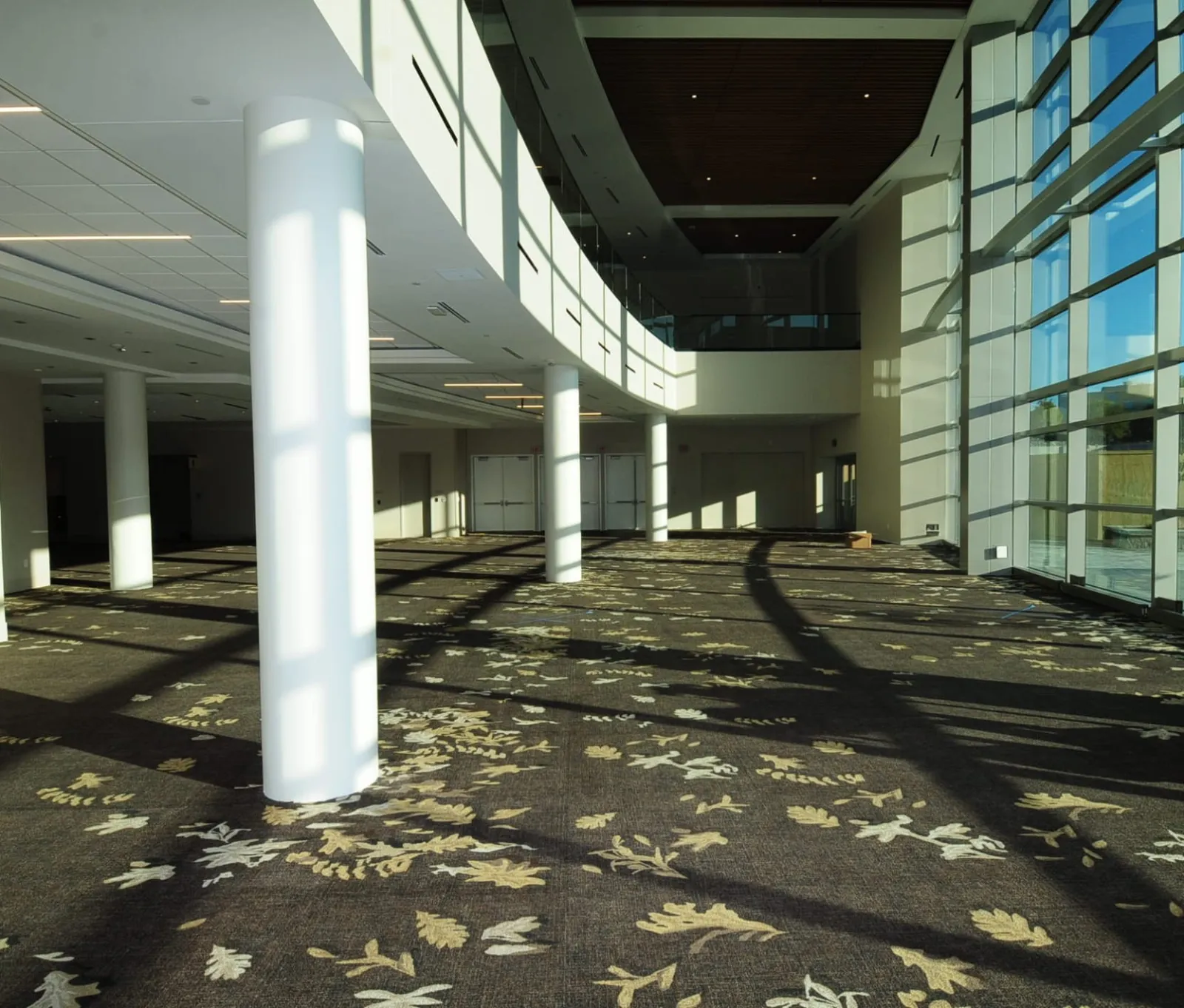 Spacious modern lobby with large windows, white columns, and patterned carpet illuminated by natural sunlight.
