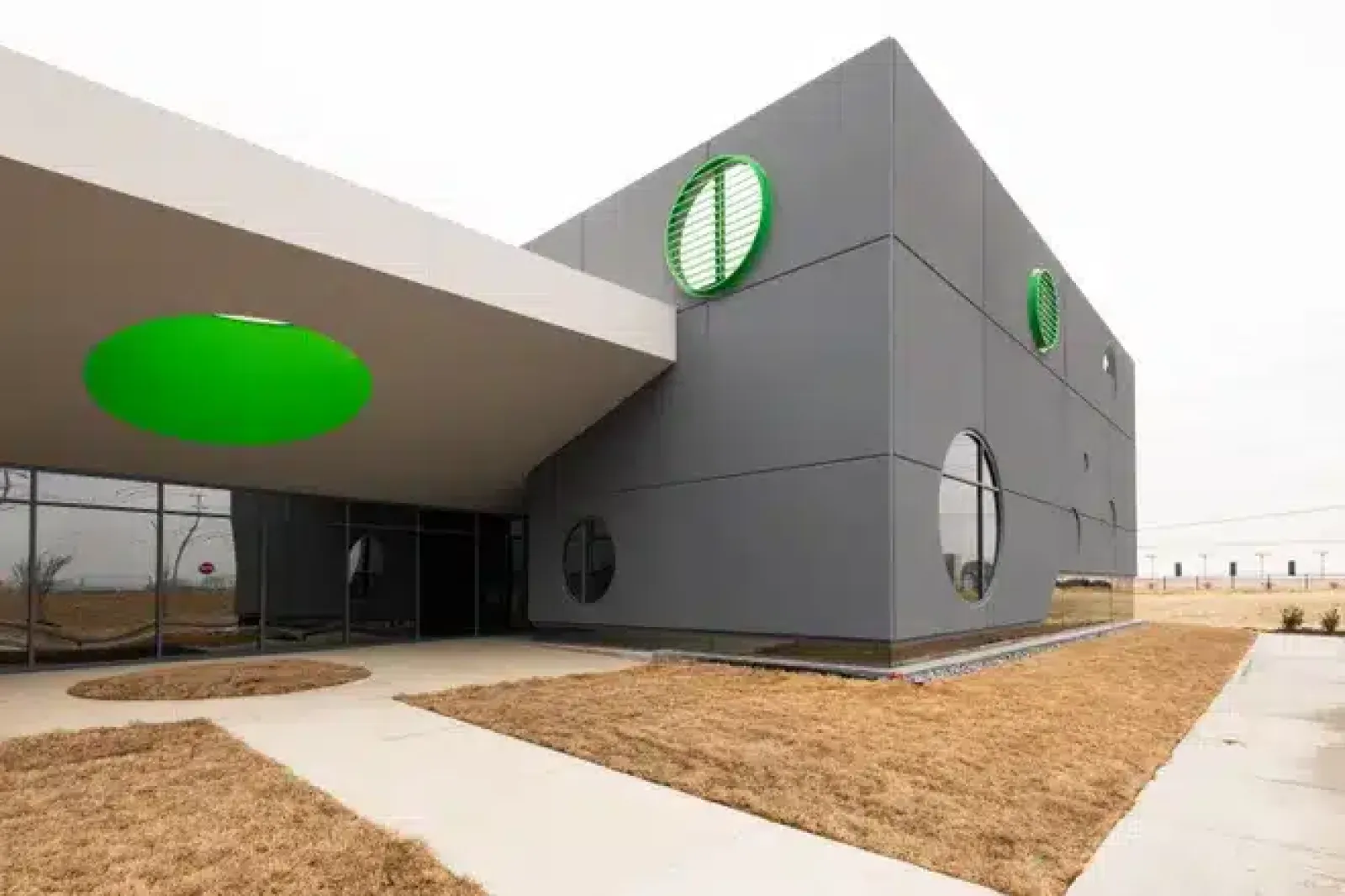 Modern gray building with circular green accents and large round windows under an overhang on dry grass landscape