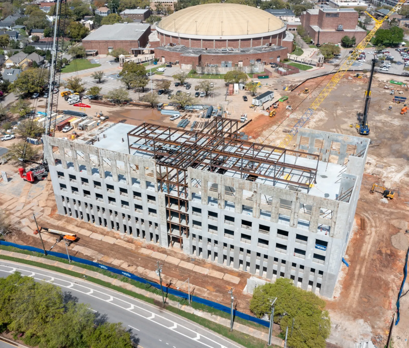 Aerial view of multi-story building under construction with steel framework and concrete walls near a domed sports arena.