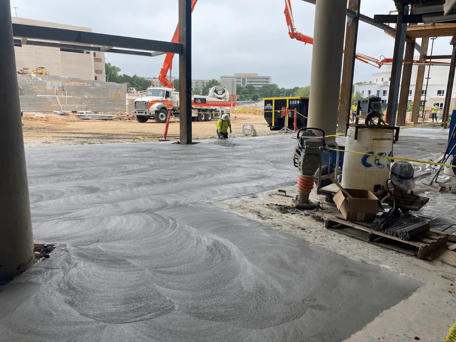 Construction site with freshly poured concrete floor and workers operating machinery under steel beams.
