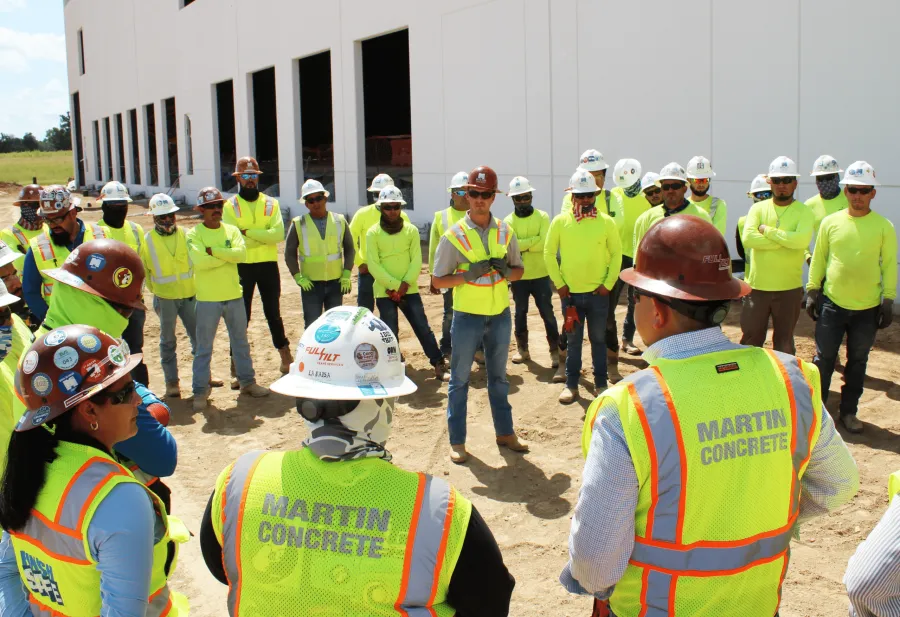 Construction workers in safety gear gather outdoors near a building site for a team meeting or briefing.