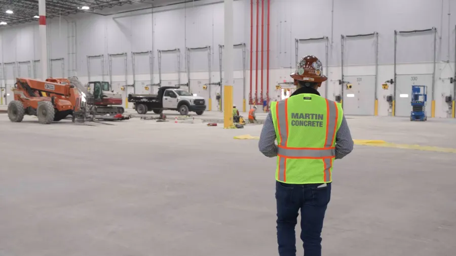 Worker in high-visibility vest with Martin Concrete logo inside large warehouse under construction.