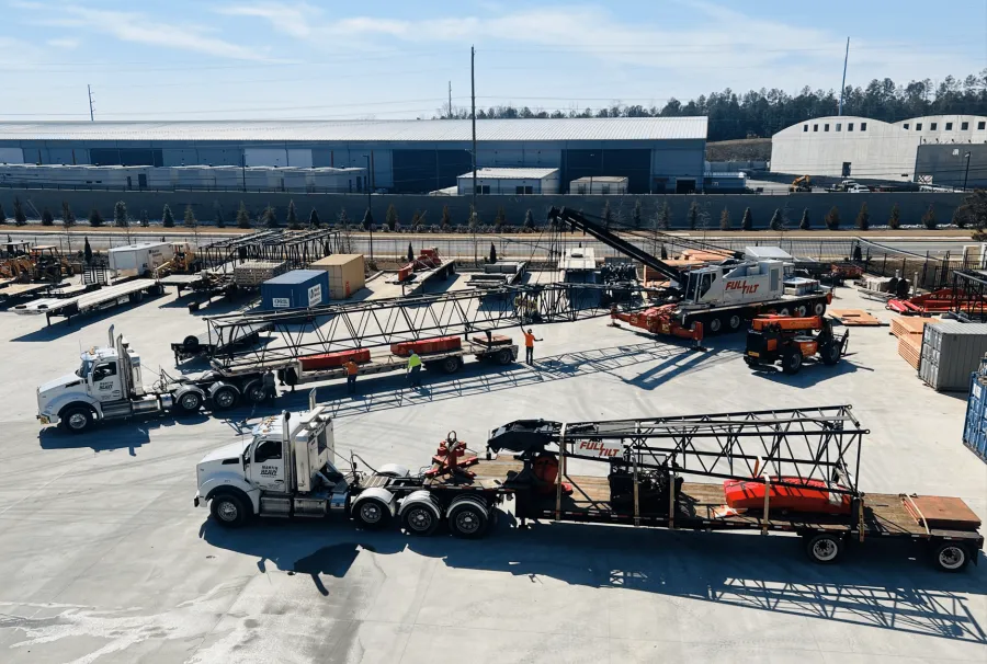 Construction site with trucks carrying large crane components and workers organizing materials under clear sky.
